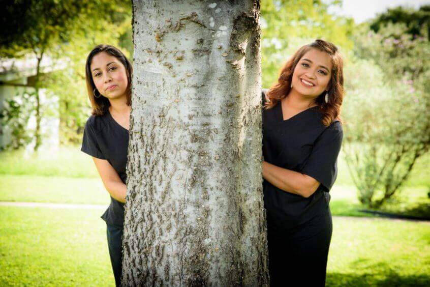 Two Crossroads Dental team members each picking from behind a different side of a big tree on the lawn in front of the dental office